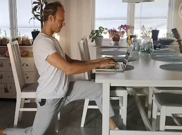 Man står på knä på golvet och sträcker ut höftböjaren samtidigt som han skriver på datorn på köksbordet.