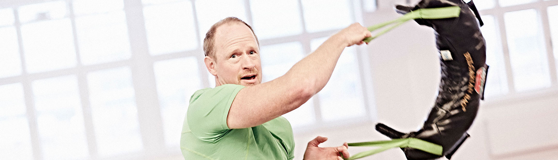 En man i grön t-shirt står i en träningssal och svingar en svart bulgarian bag åt sidan med båda händerna.