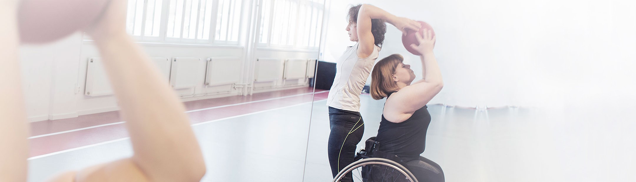 I en träningssal med spegelvägg, en stående kvinna och en kvinna som sitter i rullstol är placerade rygg mot rygg och för en boll mellan sig över bådas huvuden.