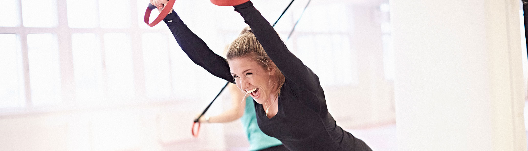 En leende kvinna i svart träningströja stäcker upp sina armar och lutar sig framåt medan hon håller händerna i ett gymband med röda handtag.