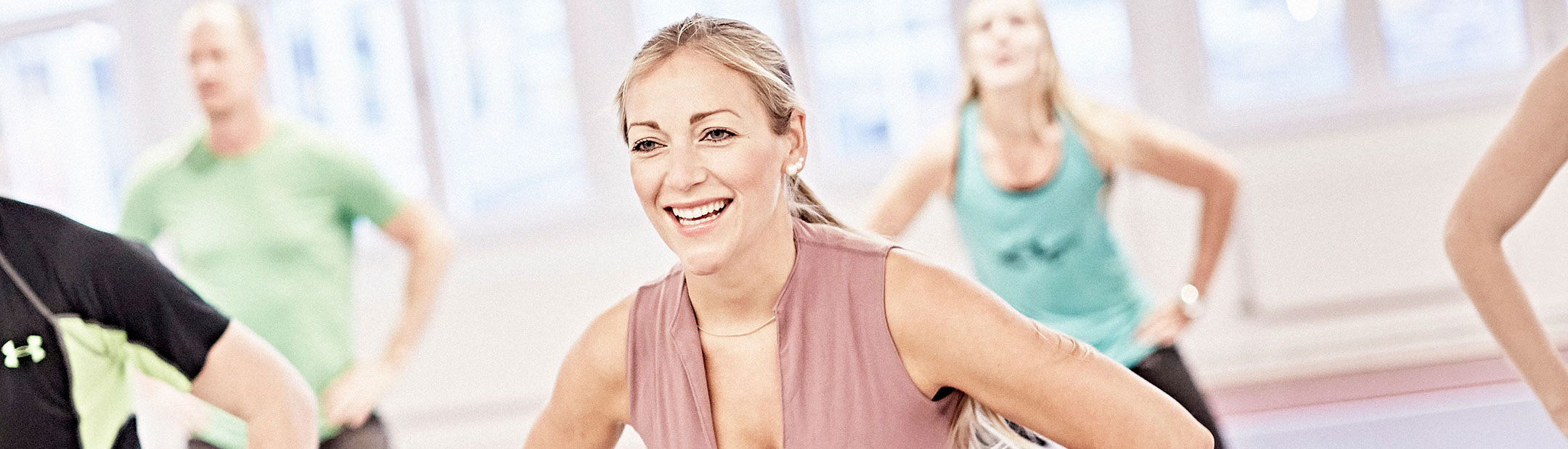 En blond, leende kvinna med rosa träningslinne är i fokus, bakom henne syns flera personer som gympar i en träningssal.