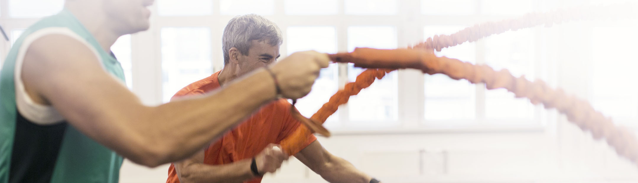 En man i grönt linne och en man i orange t-shirt står bredvid varandra och tränar med rep i en träningssal.