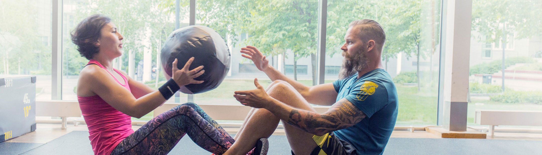 Två personer klädda i färglada träningskläder som sitter på golvet mot varandra och gör pushups med en tyngsboll